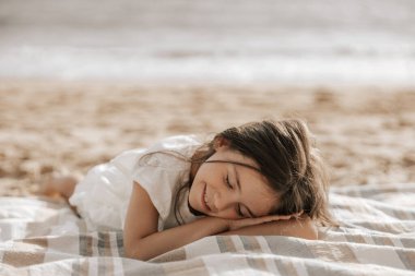 Adorable happy little girl with long brown hair in white dress smiling, while lying on blanket on sandy beach with closed eyes near wavy sea in sunlight