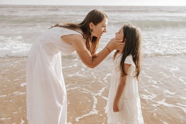 Side view of delighted young mother with long brown hair in maxi white dress, leaning forward and smiling while touching cheeks of adorable preteen daughter near wavy ocean