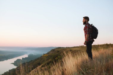 Side view of calm young bearded male hiker with dark hair in checkered shirt with backpack, standing on grassy mountain top and admiring scenic valley at sunrise