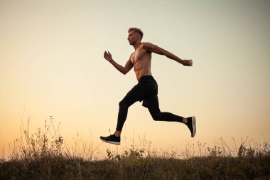 Side view of active young shirtless male athlete with muscular body jumping on grassy meadow while exercising against cloudless sunset sky in nature