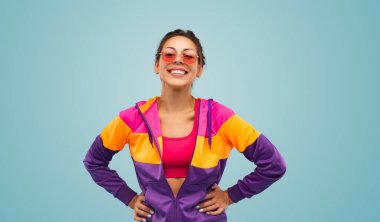 Cheerful young female athlete in multicolored hoodie and bra with sunglasses holding hands on waist, and looking at camera with smile during break in fitness training against blue background