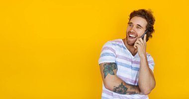 Cheerful tattooed male in striped t shirt smiling and looking away while having smartphone conversation against yellow background