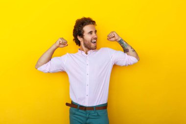 Positive tattooed young man in smart casual clothes showing biceps and looking away with smile against yellow background