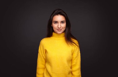 Positive hispanic young female in yellow sweater smiling and looking at camera while standing against black background