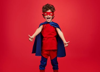 Happy little boy in superhero costume screaming and gesturing thumbs up against red background