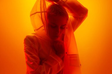 Stylish female model with short blond hair adjusting hood of futuristic raincoat and looking down thoughtfully in bright orange studio with neon light