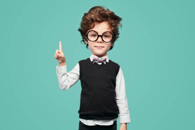 Cute clever little schoolboy with big had and brown hair in classy clothes with bow tie and round eyeglasses, pointing up and looking at camera against turquoise background