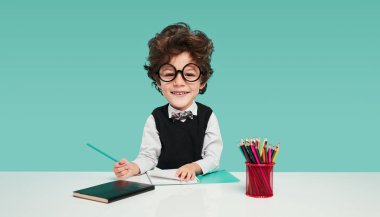 Cute boy in school uniform and glasses with big head smiling and looking at camera, while sitting at desk and doing homework assignment against turquoise background