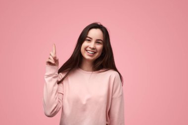 Happy female teenager in sweatshirt with long dark hair looking at camera with smile and pointing up during advertisement campaign against pink background