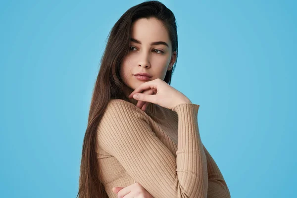 Teenager in beige turtleneck with long dark hair looking away and thinking while touching chin against blue background