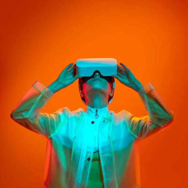 Young female in transparent futuristic coat touching VR headset and looking up while exploring cyberspace under neon light against orange background.