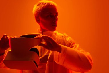 Self assured young futuristic woman with short hair in jacket holding modern VR headset and looking away against bright orange background
