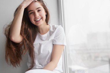 Cheerful teen brunette looking at camera with smile and touching long hair while resting near window at hoe