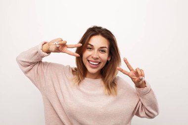Confident young female model with brown hair in sweater smiling brightly and showing two fingers gesture against white background