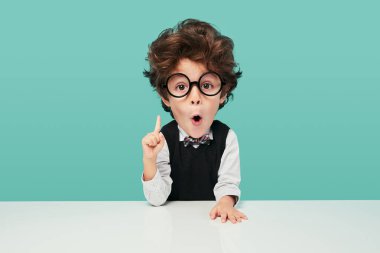 Funny little boy in school uniform and glasses with big head looking at camera with opened mouth, and pointing up while sitting at desk during lesson against turquoise background