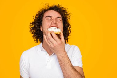 Happy young hungry man with curly dark hair in polo shirt eating delicious glazed sweet doughnut with closed eyes against yellow background