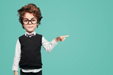 Adorable smart little boy with dark hair and big head in formal outfit and round eyeglasses, pointing aside and looking at camera against turquoise background