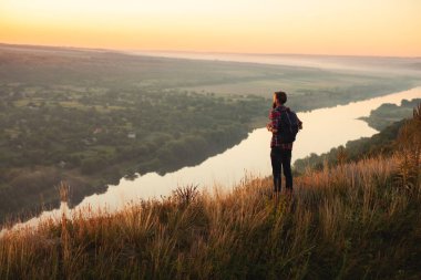 Man in casual clothes with backpack standing on grassy hill and observing calm river during trip in countryside at sundown