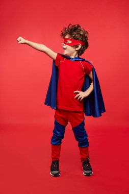 Full body funny boy in superhero costume holding hand on waist and clenching fist while standing against red background