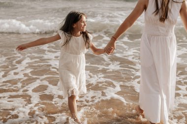 Crop unrecognizable young woman with long dark hair in maxi white dress holding hand of positive little daughter, while walking together in sea water on sandy beach