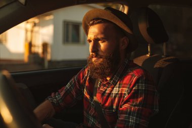 Bearded male hipster in checkered shirt and hat sitting on passenger seat of vehicle during ride in morning in city
