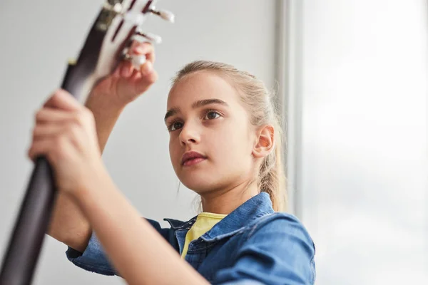 Low Angle Cute Girl Turning Pegs Guitar While Standing Window — Foto Stock
