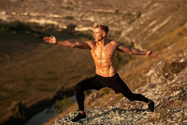 Full body of young shirtless male athlete with muscular naked torso performing Warrior II pose, while practicing yoga on edge of rocky cliff in mountainous valley