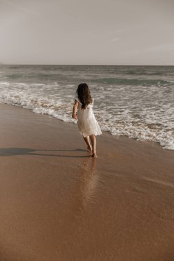 Back view of unrecognizable barefooted girl with long dark hair in white dress running on wet sandy beach near foamy sea during summer holidays