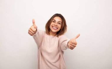 Excited young female model with brown hair in casual clothes smiling happily and looking at camera while showing thumbs up gesture against white background