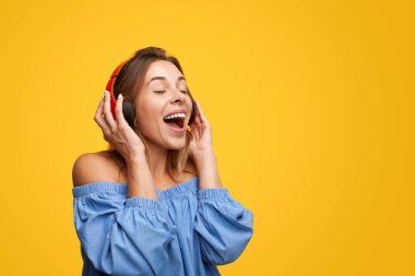 Optimistic young female with long brown hair in blue blouse with bare shoulders, singing happily with closed eyes while listening to favorite song in wireless headphones against yellow background