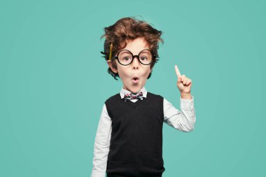 Adorable little boy in school uniform and glasses looking at camera and pointing up while having idea against turquoise background