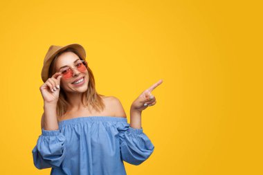 Self assured young female millennial with long dark hair in stylish outfit and sunglasses smiling happily and pointing away against yellow background