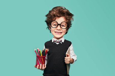 Funny clever little boy with big head in classy outfit and round spectacles smiling happily and looking at camera, while standing against turquoise background with copybook and colorful pencils