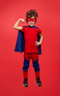 Full body of positive little boy in superhero costume with cape and mask smiling and looking at camera while showing bicep against red background