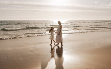 Full body of young barefooted woman with long hair in maxi white dress holding hand of adorable little daughter, while walking together along wet sandy beach near wavy ocean