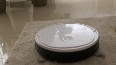 White round robot vacuum cleaner cleaning carpet floor. Following shot of home autonomous smart bot vacuuming apartment by itself.