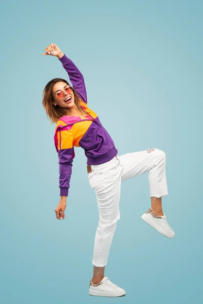 Side view of positive young cool female millennial with long hair in trendy outfit and sunglasses, smiling while standing against blue background with raised arm and leg