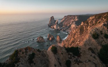 Breathtaking drone view of rough rocky cliffs of Cape Roca located near waving sea against sunrise sky in morning in Portugal