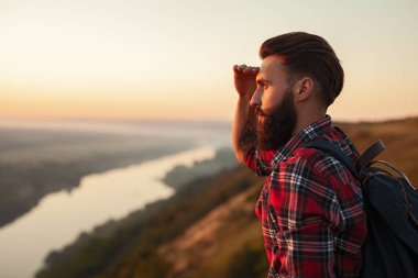 Side view of bearded man in checkered shirt with backpack touching forehead and looking away while admiring countryside at sunset