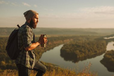 Side view of young bearded male traveler in checkered shirt with backpack drinking hot tea, from thermos and admiring scenic landscape of river and valley during hiking trip