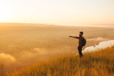Side view of young bearded male traveler in casual clothes and hat with backpack, pointing away while standing on grassy slope of mountain during hiking trip at sunrise