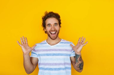 Merry young man in striped t shirt raising arms and yelling in excitement against yellow background