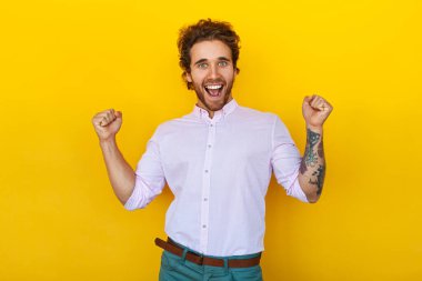 Cheerful young man in smart casual clothes with tattooed arm clenching fists and screaming while celebrating victory against yellow background