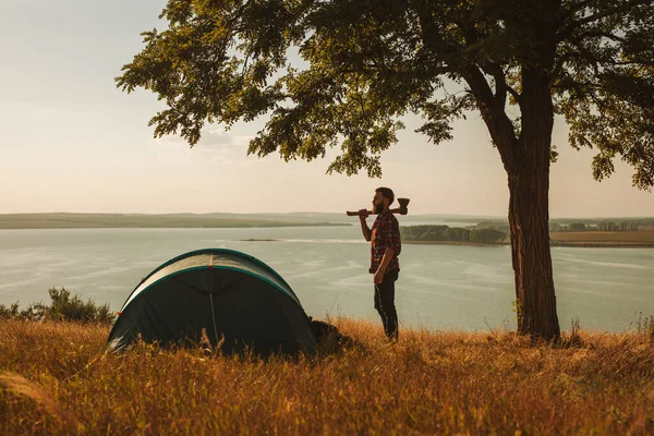 Side view of young bearded male hiker in checkered shirt standing with axe in hand, on grassy meadow near tent placed at lakeside during trip in countryside
