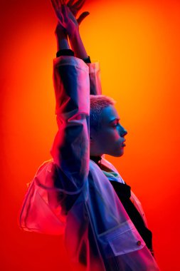 Side view of young androgynous model in futuristic outfit raising arms and looking away under colorful neon illumination against orange background
