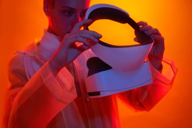 Crop focused female in transparent coat putting on modern VR goggles while standing in bright studio with neon lights before exploring cyberspace