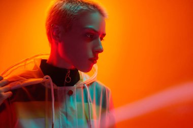 Serious young female millennial with short blond hair in transparent raincoat touching hood and looking away against orange background in studio with neon lights