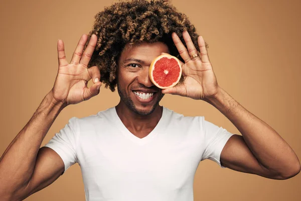 Cheerful African American man in white t shirt with curly hair holding half of fresh grapefruit near eye and gesturing OK against brown background