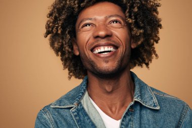 Happy young black male in denim jacket with curly hair looking up and laughing against brown background