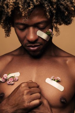 Vulnerable shirtless African American male with fresh patches attached to damaged skin with patches clasping hands on chest against brown background